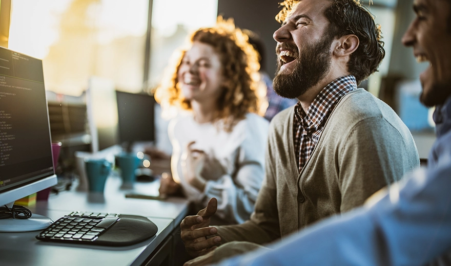 Three people sitting in front of a desk looking towards a computer monitor and laughing.