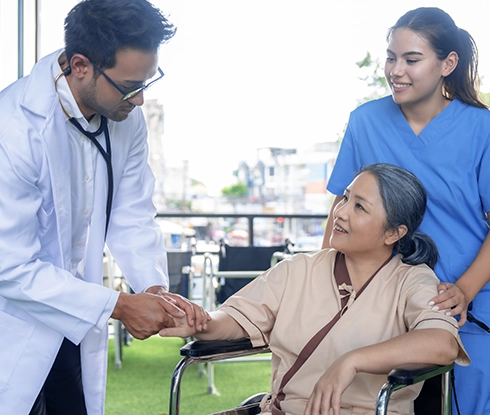 A doctor checks the pulse of a patient in a wheelchair. Behind the patient is a medical assistant wearing scrubs