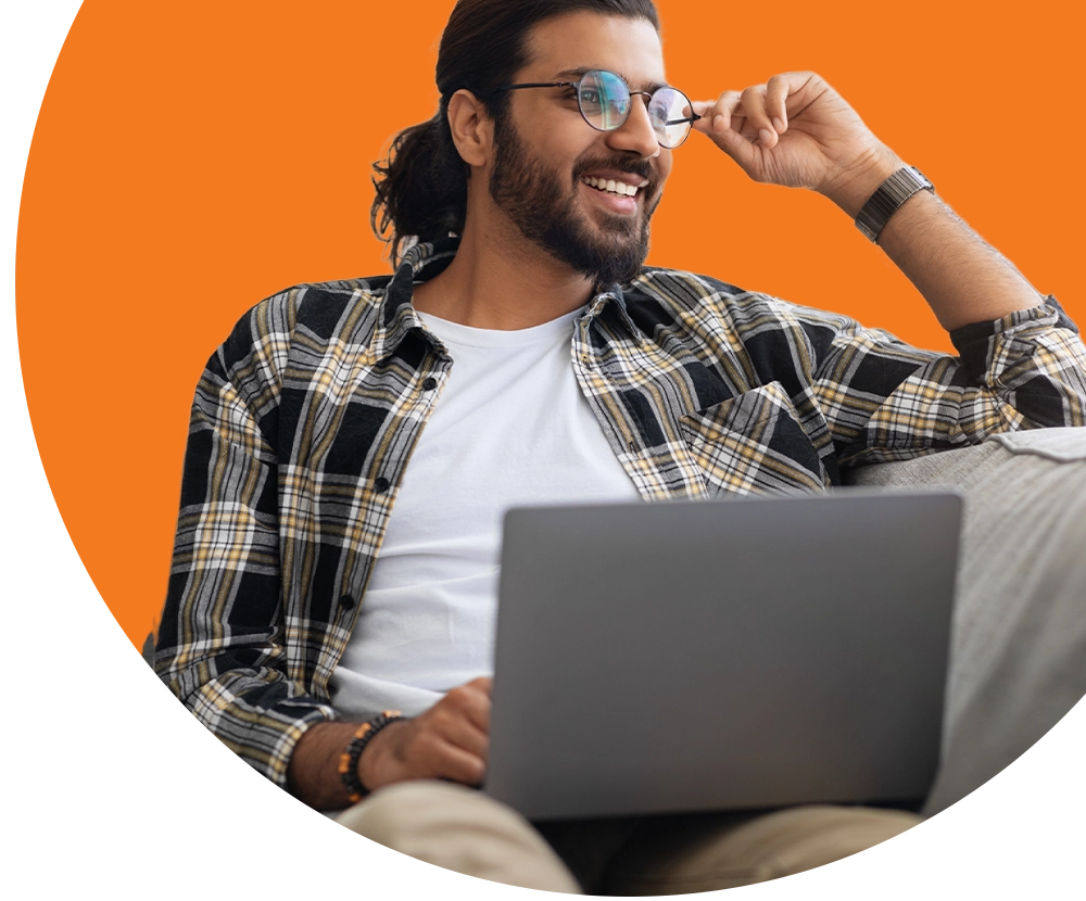 A man wearing glasses and flannel sits on a couch working on a laptop