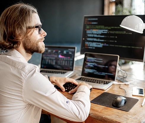 A man writing code. In front of him are a desktop monitor and two laptops